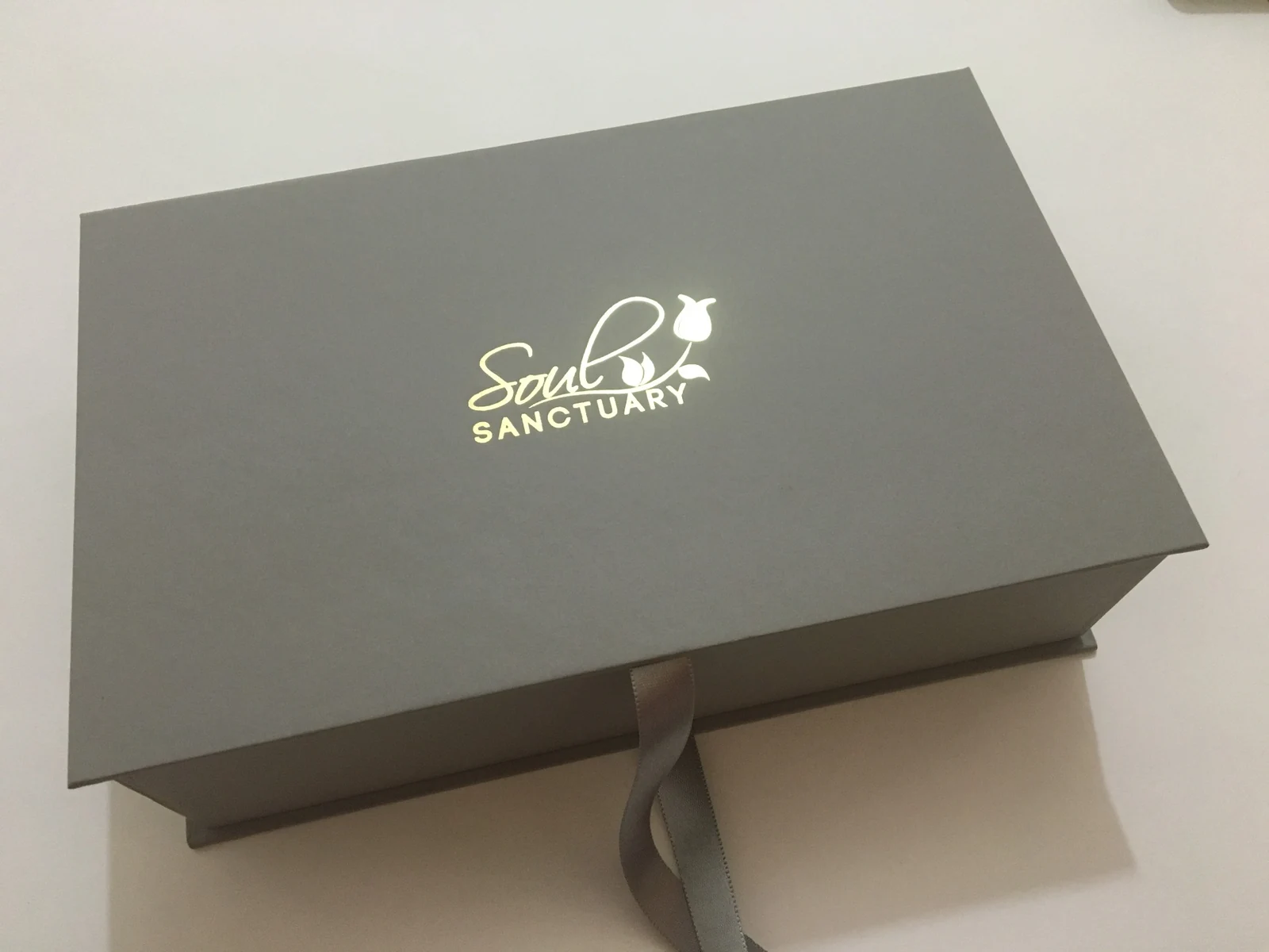 Grey rectangular presentation box with gold branding and a ribbon closure